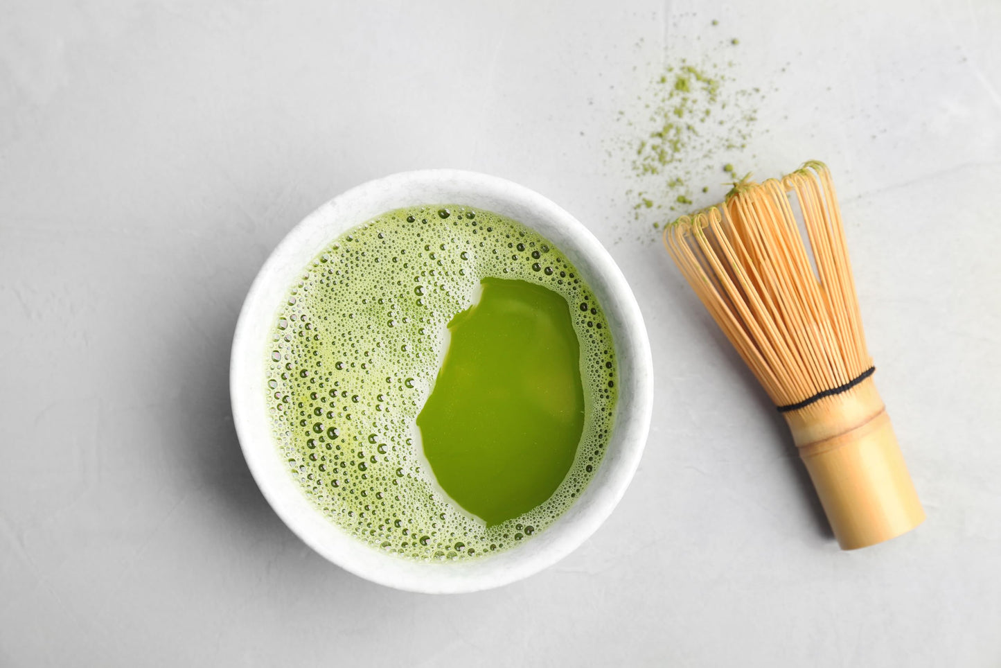Matcha green tea in a white bowl with a bamboo whisk on a light grey background