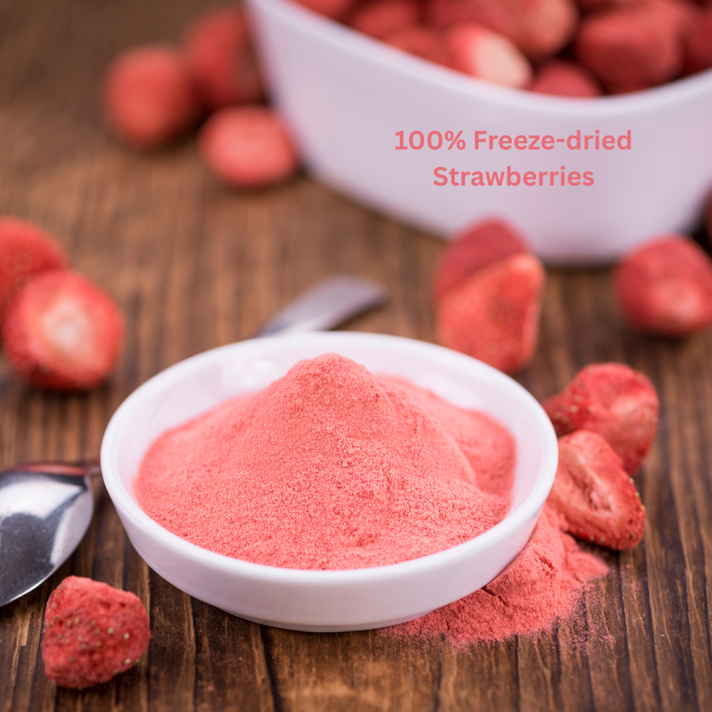A close-up, slightly elevated view of a small white bowl filled with finely ground pink strawberry powder, resting on a brown wooden surface. Freeze-dried strawberry pieces and a silver spoon are scattered around the bowl. A larger, out-of-focus white bowl of freeze-dried strawberries is visible in the background. The text "100% Freeze-dried Strawberries" is written in the upper-right area.